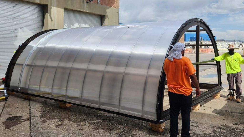 Two workers, one in an orange shirt and the other in neon green, stand beside a large, transparent barrel vault skylight structure outdoors on a sunny day.