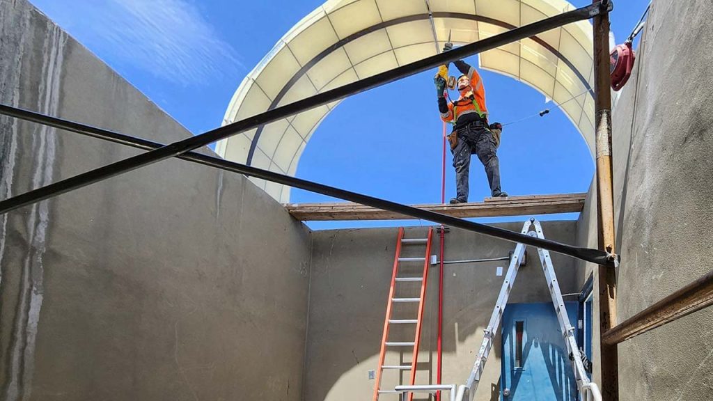 A construction worker in an orange vest and hard hat stands on scaffolding removingfiberglass skylight panels, surrounded by ladders and blue sky, suggesting focus and safety.