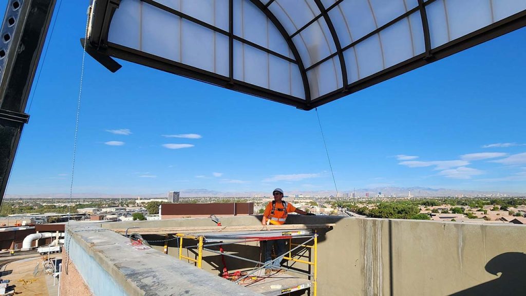 A construction worker in a safety vest stands on a rooftop scaffold as a crane llowers the new skylight against a clear blue sky, overlooking a city skyline.