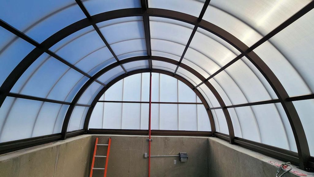 Interior of a polycarbonate barrel vault skylight with metal framework, creating a bright and airy space. An orange ladder rests against a gray concrete wall.