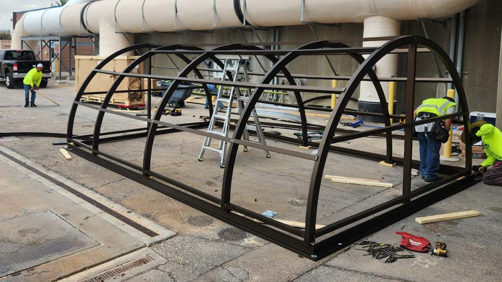 Workers in neon vests construct a large metal barrel vault skylight frame with arched supports on a concrete surface. A ladder and tools are scattered nearby, indicating active construction.
