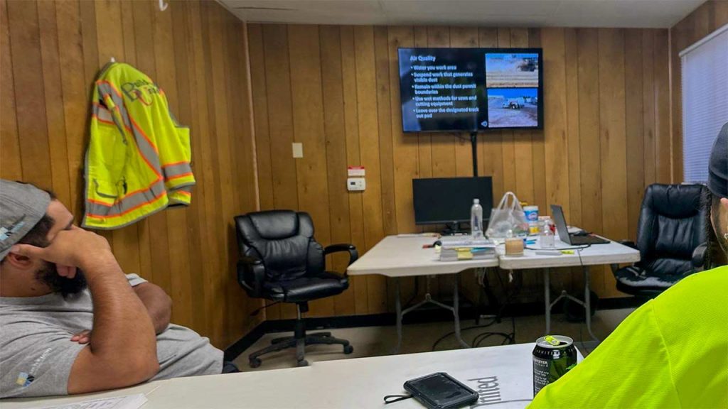Two technicians sit at a conference table in a wood-paneled room. A wall-mounted screen displays a slide about air quality. The setting conveys a professional, focused atmosphere.