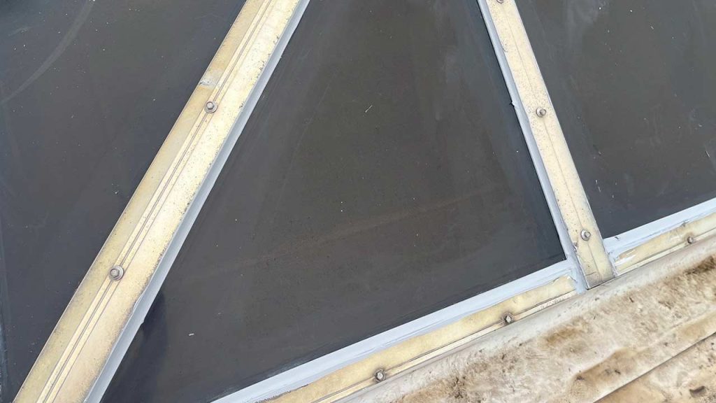 Close-up of a triangular glass pane with a dark surface, bordered by beige metal strips and rivets. A textured, weathered area is at the bottom.