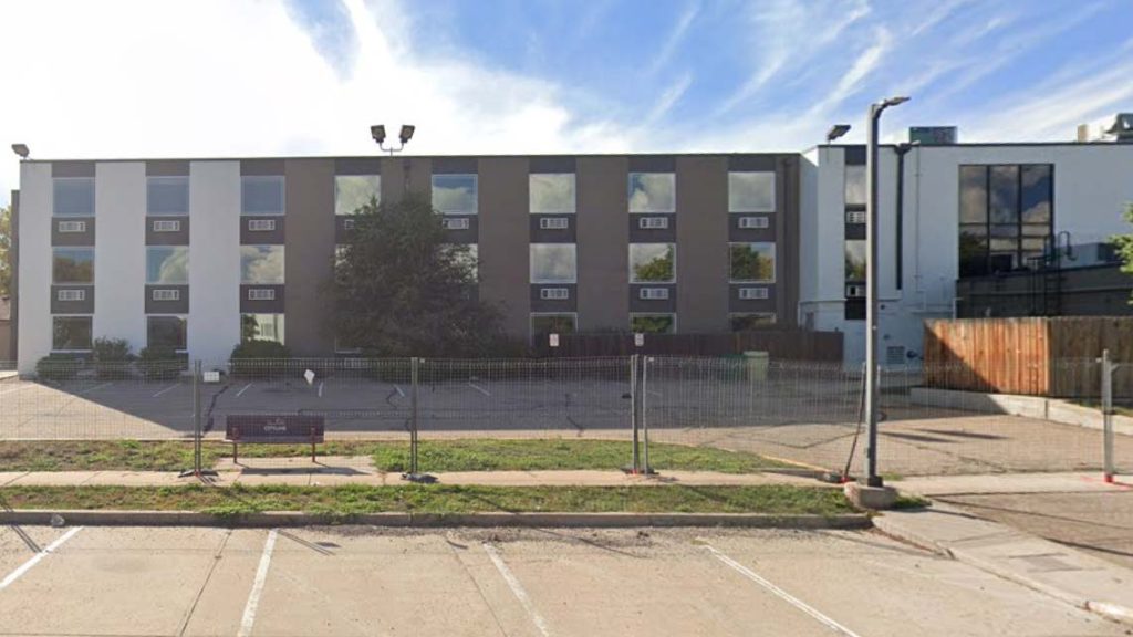 A three-story building with a mix of white and gray facade sits behind a fenced parking lot under a bright blue sky with clouds. The scene is calm.