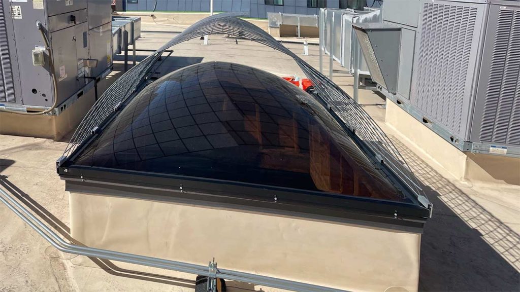 A domeskylight on a rooftop, surrounded by security mesh netting for protection. The structure is flanked by HVAC units, creating an industrial feel.