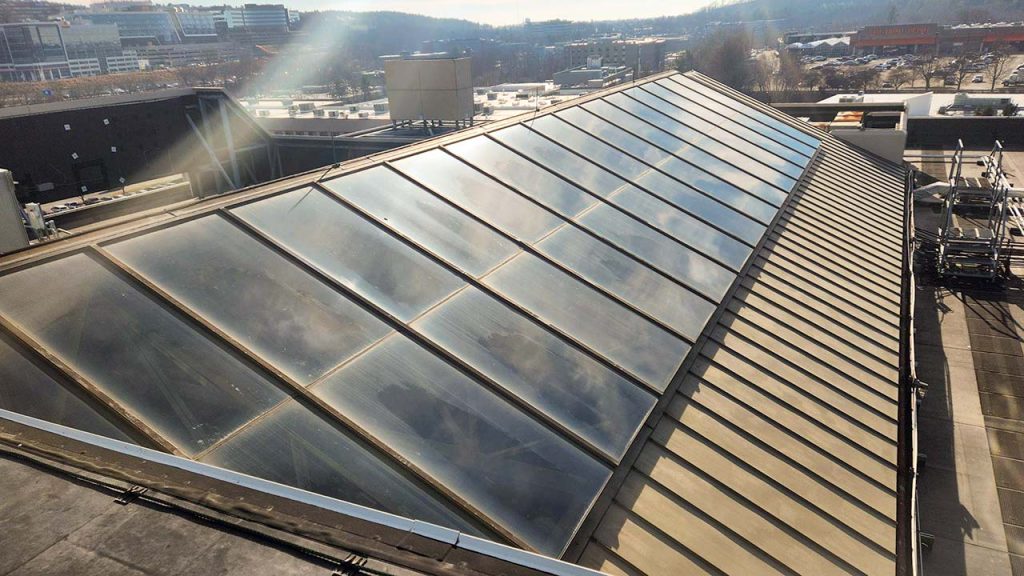 Rooftop atrium skylight with large glass panels reflecting sunlight, set in an urban industrial area. Background shows buildings and distant hills under a clear sky.