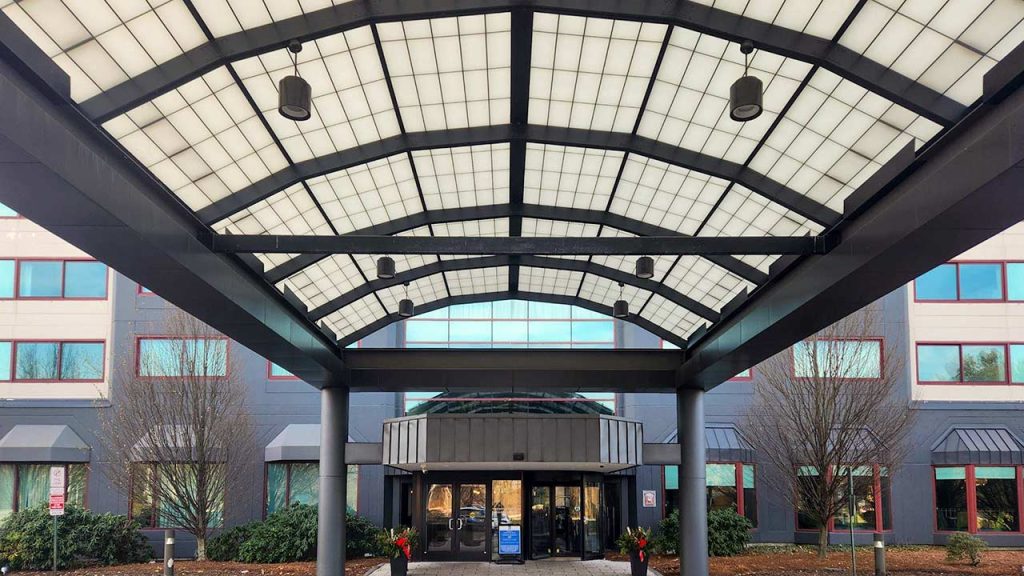 The image shows the entrance of a modern building with a large, arched fiberglass canopy. The building has grey walls and red-framed windows. The scene is calm and orderly.