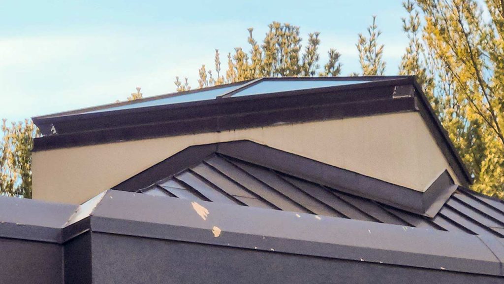 A metal roof with a pyramid skylight shows peeling paint on a modern building under a clear blue sky. Tall trees surround the structure, creating a serene atmosphere.