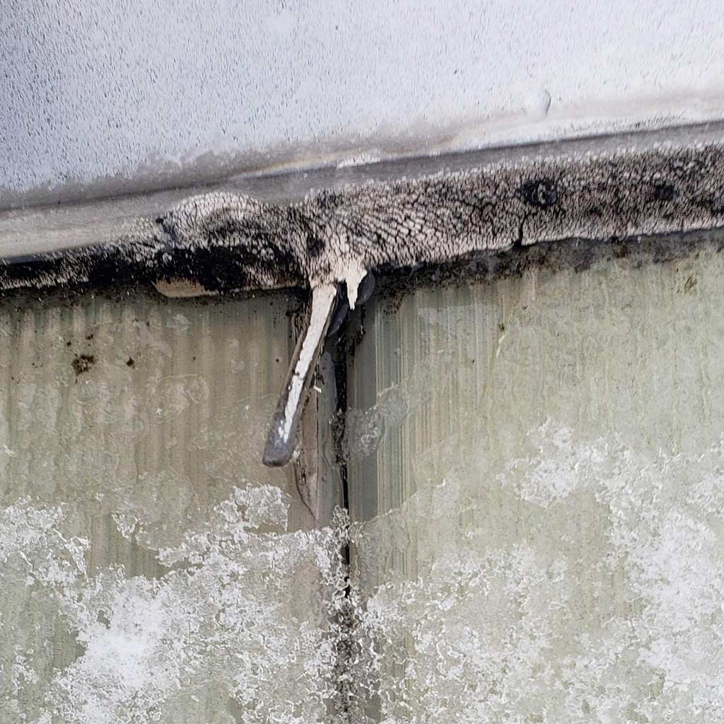 A small fish, piece of caulking hangs from a metal surface covered in frost. The texture is rough, with visible ice crystals, conveying a cold and harsh atmosphere.