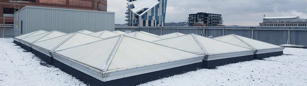 Snow-covered rooftop with a cluster of , pyramid skylights. A gray, overcast sky and urban buildings in the background convey a cold, wintry atmosphere.