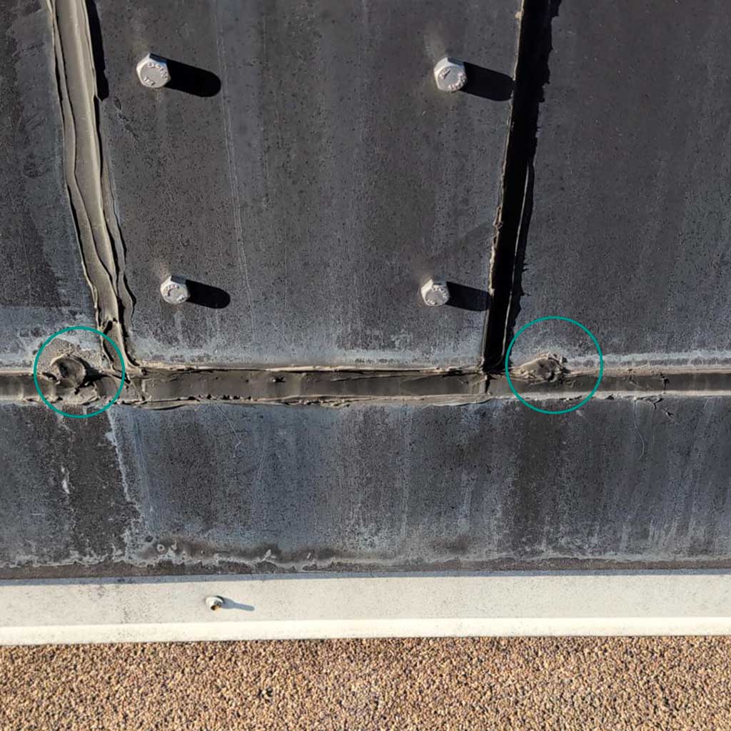 Close-up of a weathered metal surface with visible bolts and two plugged weep holes circled in green, suggesting wear or damage. The tone is industrial and somber.