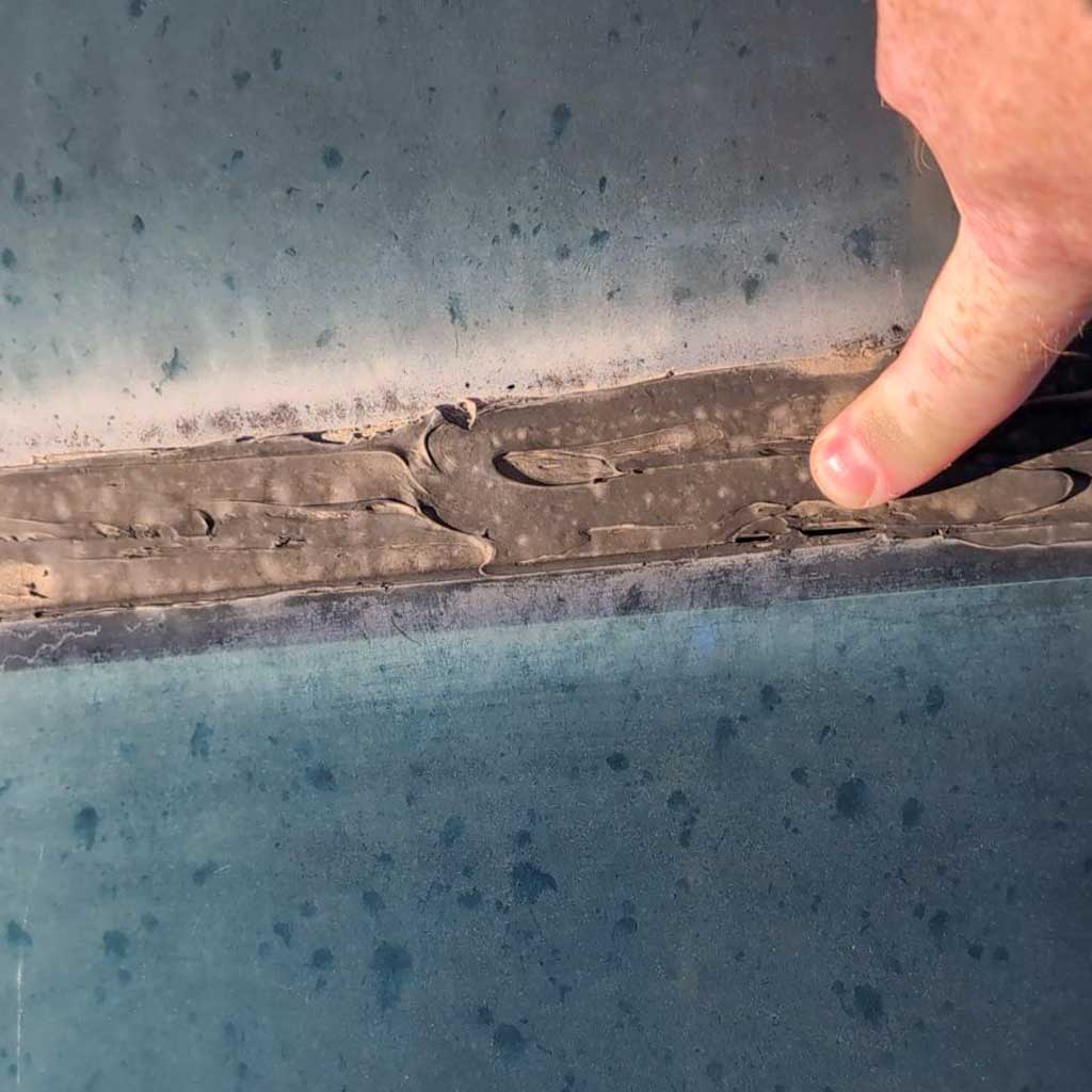 A hand points at a cracked, peeling seam of caulking on a dirty blue skylighjt glass. The texture is rough and uneven, highlighting wear and age. The mood suggests neglect.