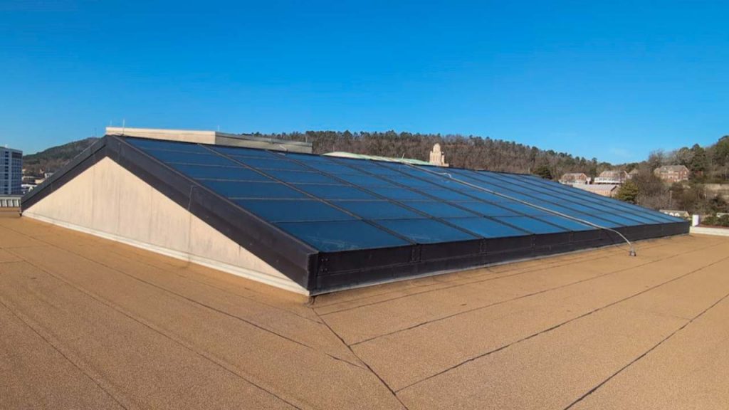 A large, modern skylight with dark-tinted glass panels sits on a flat rooftop against a clear blue sky. Surrounding hills and buildings are in the background.