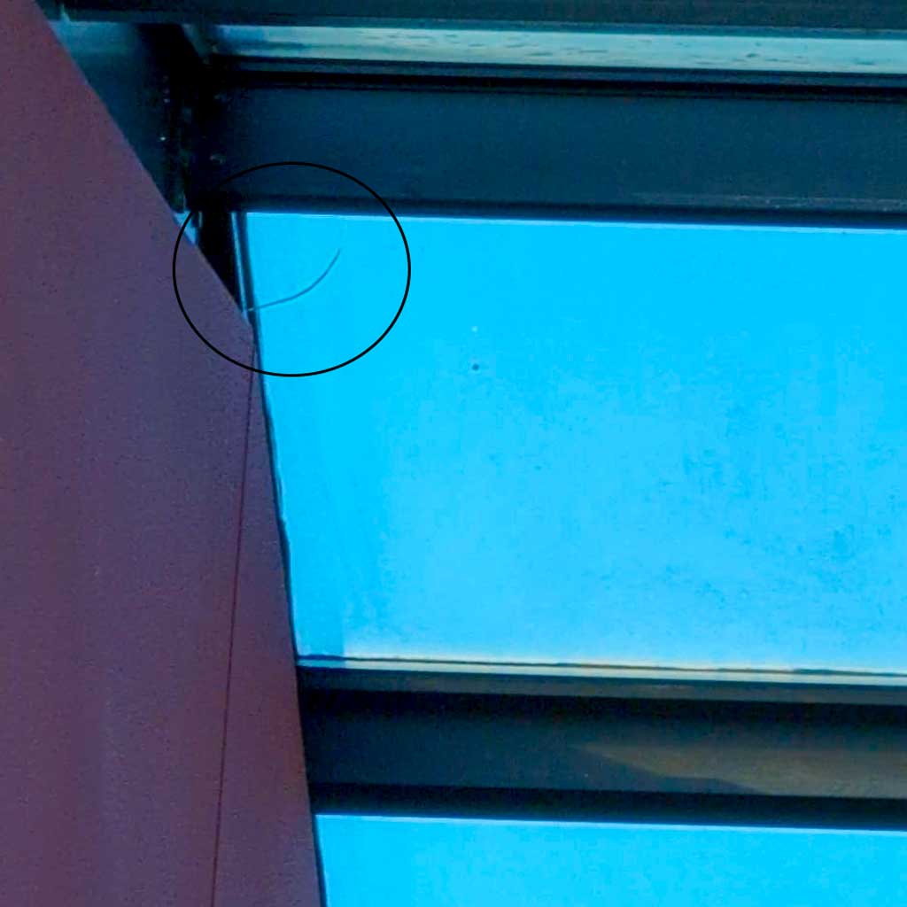Interior view of a skylight corner with a metal beam against a vibrant blue sky. A small crack is circled near the top, highlighting potential damage.