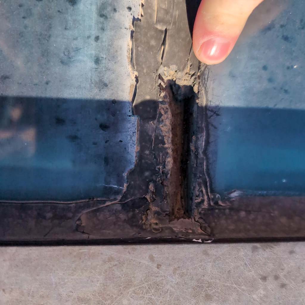 Close-up of a finger pointing at cracked, peeling caulking on a weathered skylight seam, revealing a gap underneath. The image conveys neglect and decay.