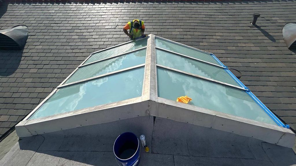 A worker in a safety vest cleans a large glass skylight on a sloped roof, reflecting blue sky and clouds. Supplies are next to the skylight.