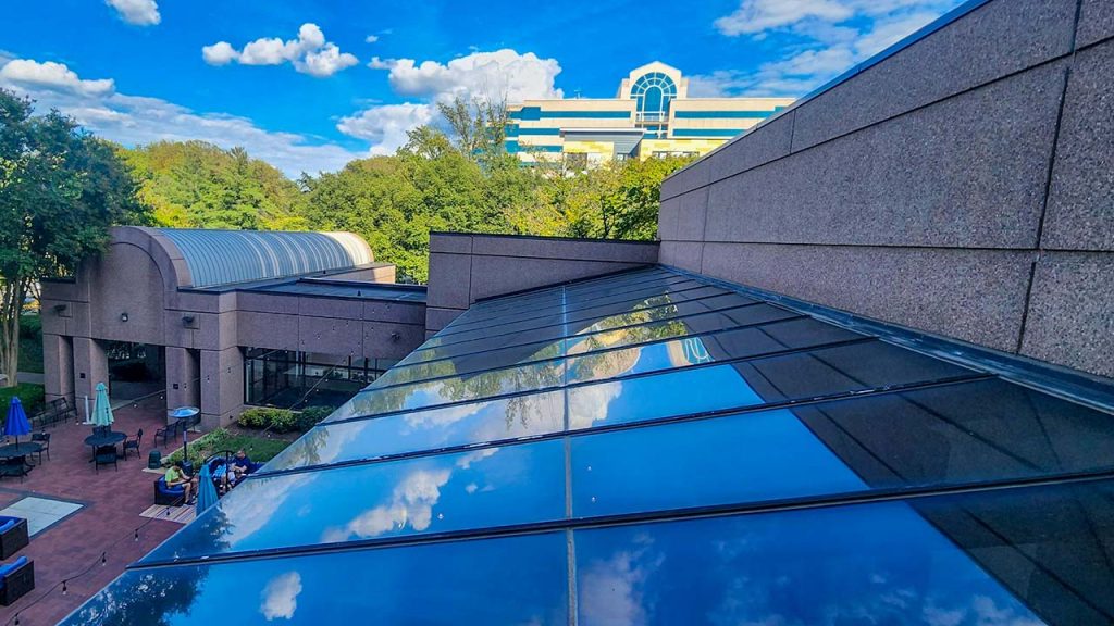 Modern building with glass skylight reflecting a vibrant blue sky and fluffy clouds. Surrounded by lush green trees, creating a serene atmosphere.