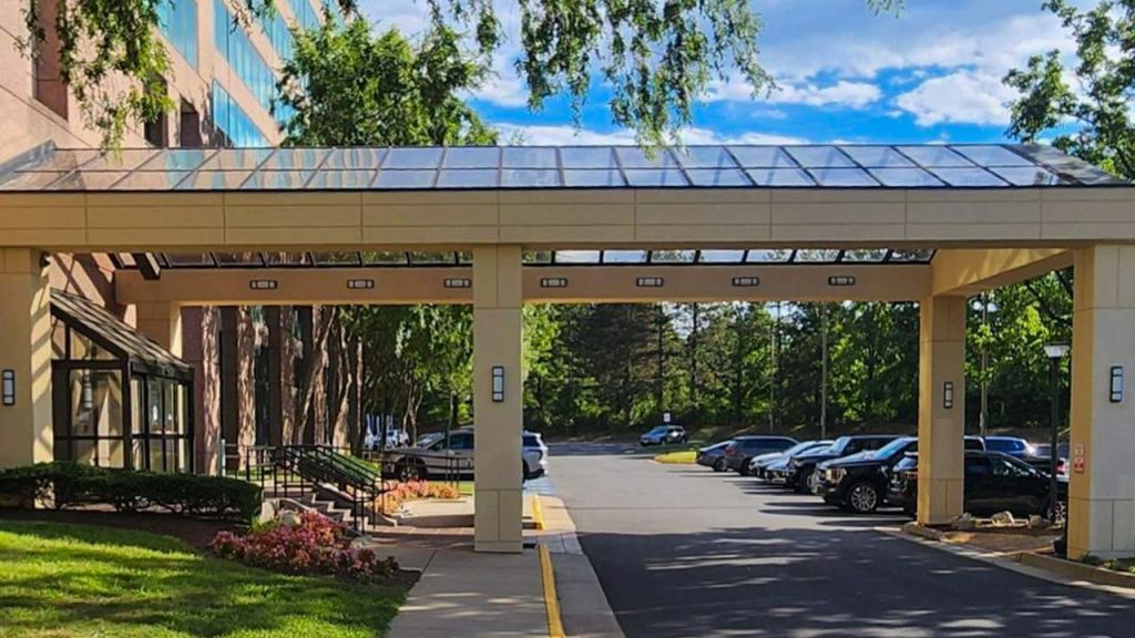 A covered walkway leads to a building entrance surrounded by lush greenery and parked cars under a bright blue sky, creating a welcoming atmosphere.