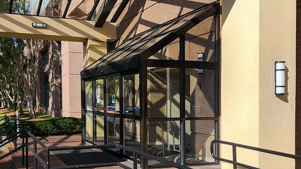 Modern building entrance with glass doors and metal frame, flanked by beige walls. Shadows from overhead structure create a geometric pattern.