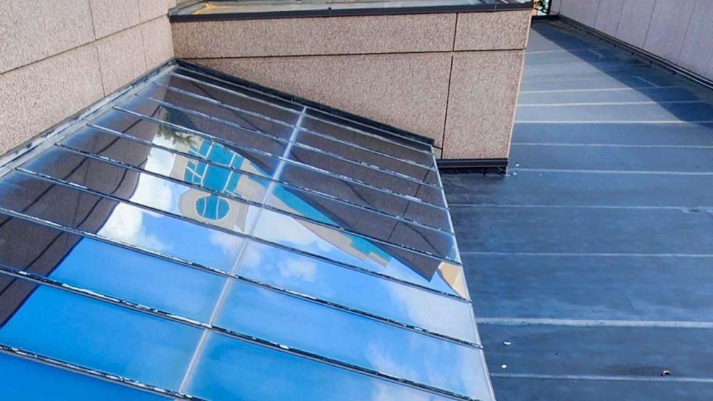 Rooftop view showing a modern building's glass single slop[e skylight reflecting blue sky and clouds, blending sleek architecture with serene outdoor elements.