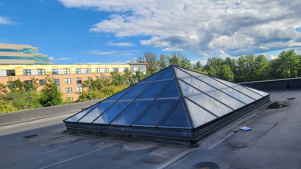 A glass pyramid skylight on a flat rooftop reflects the cloudy blue sky. In the background, there are trees and a multi-story building.