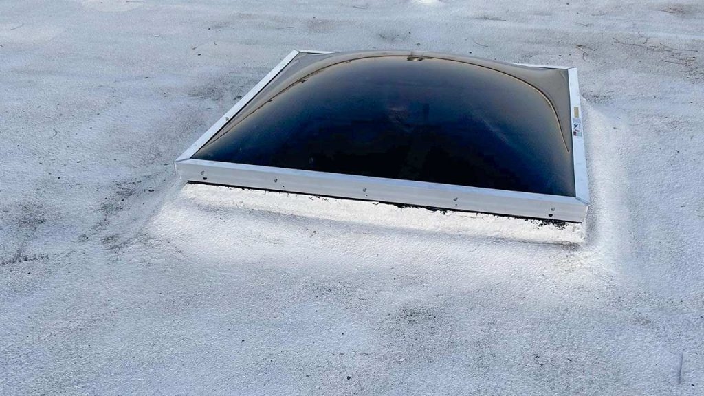 A dome-shaped, dark-tinted skylight set in a white, textured flat roof. The skylight contrasts with the bright roofing, giving a modern and sleek appearance.