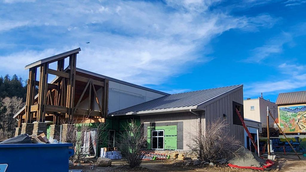 Partially constructed building with wood framing, gray siding, and exposed insulation. Bright blue sky and scattered clouds create a hopeful tone.