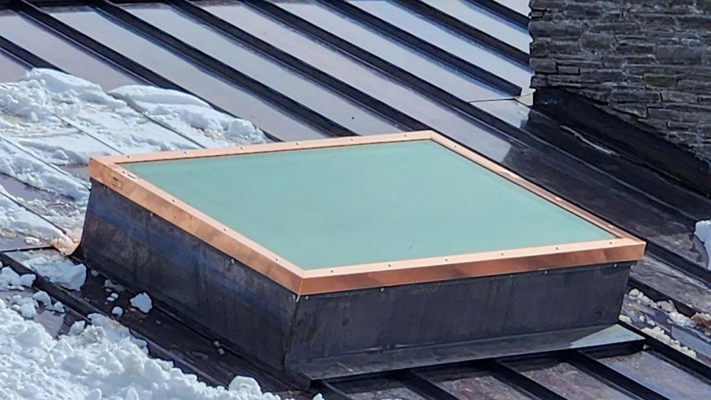 A square skylight on a metal roof surrounded by snow. The skylight has a copper frame and is set against a backdrop of stone wall and roofing panels.