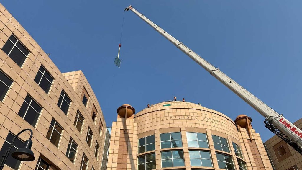 A crane hoists a glass pane over a modern, beige stone building under a clear blue sky. The structure features large windows and a curved facade.