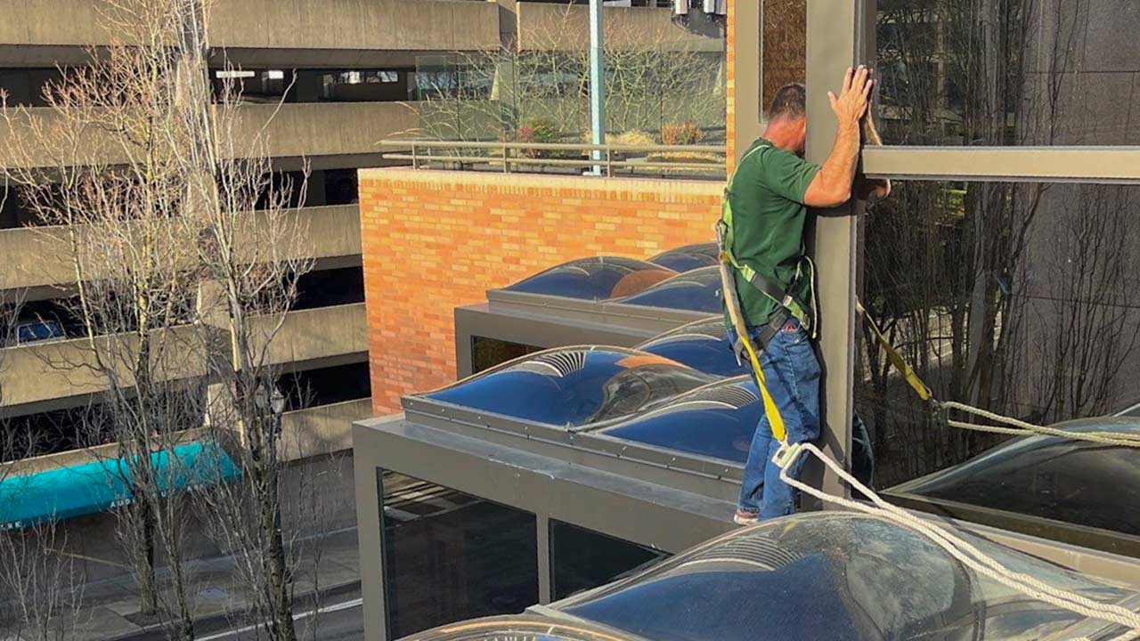 A man in a green shirt is precariously walking on a narrow ledge of a building, wearing a safety harness. He grips a large glass window. Urban background.