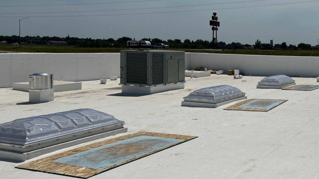 Flat rooftop with white surface, featuring skylights, HVAC unit, and air vents. Background shows distant highway and signage under a clear sky.