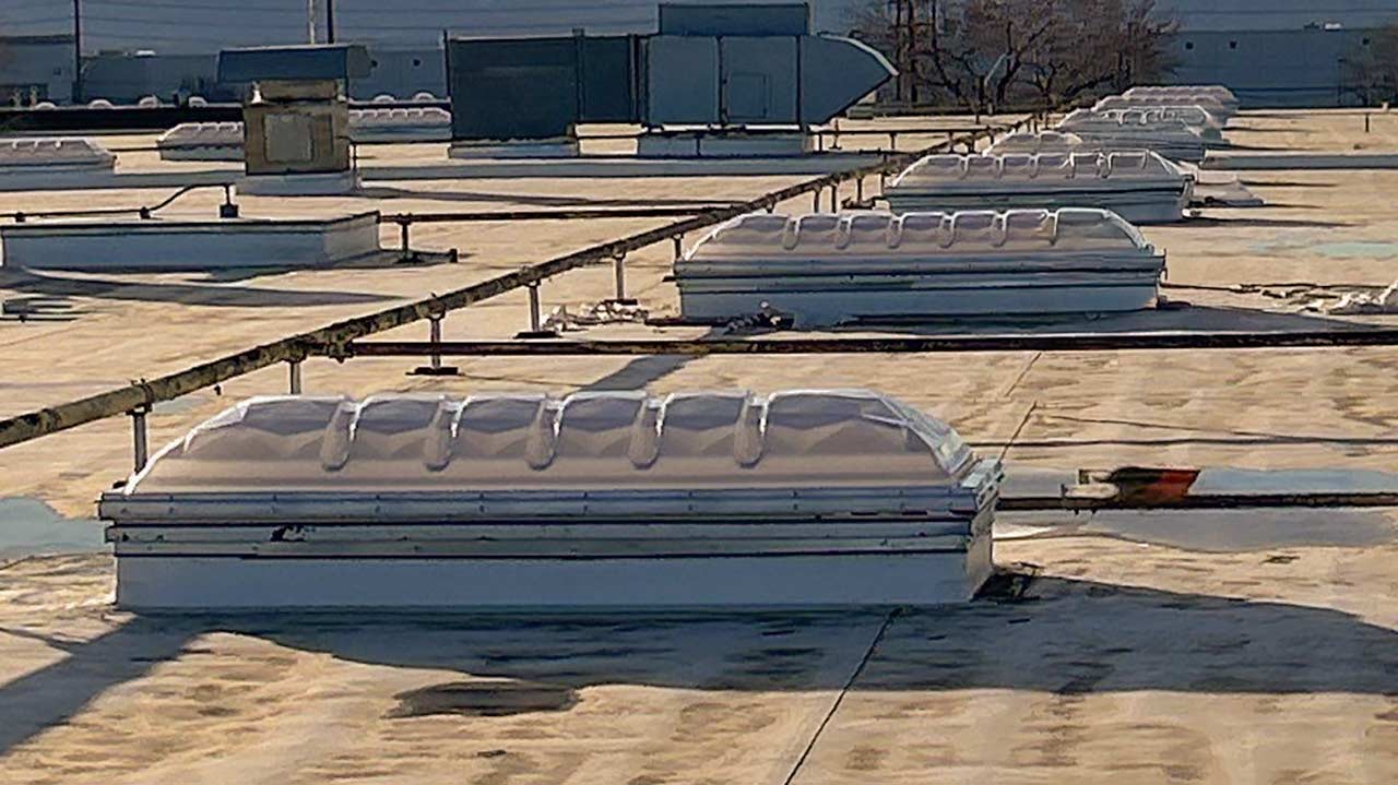 Flat industrial rooftop with multiple white skylights aligned in rows. Sunlight casts shadows, creating a geometric pattern and a calm atmosphere.