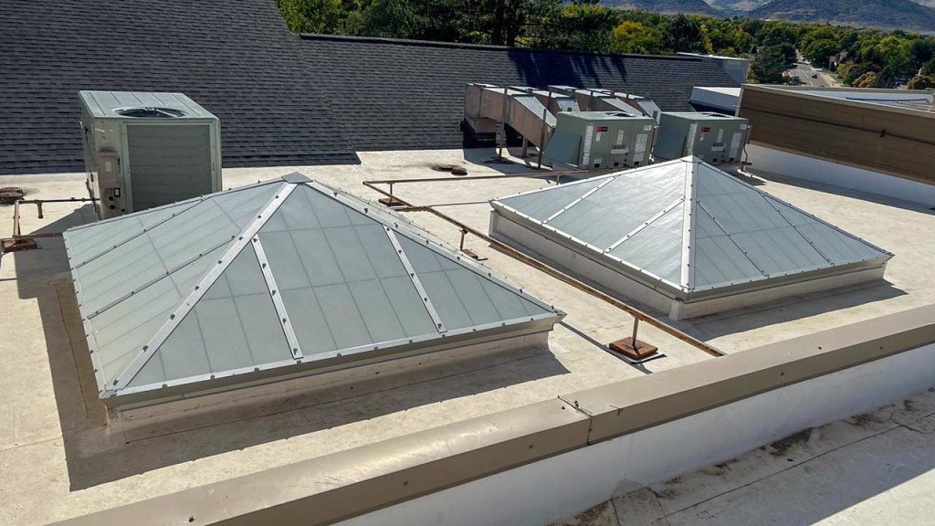 Flat rooftop with two pyramid-shaped skylights and several HVAC units against a clear sky, surrounded by trees and hills. Clean, industrial look.
