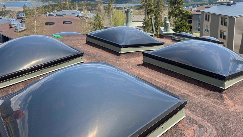 Rooftop with four dome skylights on a brown roof, surrounded by trees and buildings under a clear blue sky, creating a calm, contemporary scene.
