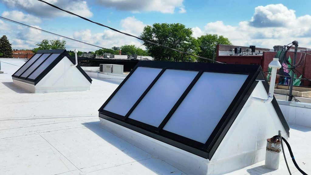 Two ridge skylights on a flat white rooftop under a blue sky with clouds. Trees and buildings are visible in the background, creating an urban setting.