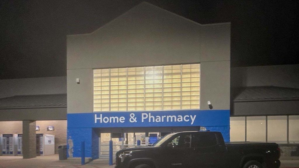 Nighttime view of a store entrance with wall panels labeled "Home & Pharmacy." A dark pickup truck is parked in front. The scene is quiet and dimly lit.