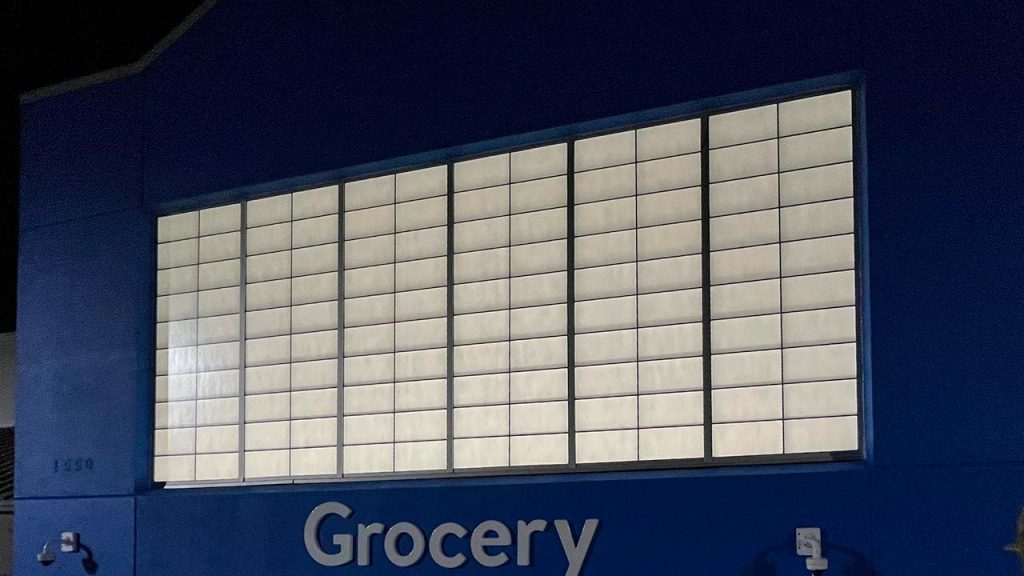 Exterior of a grocery store at night with a large illuminated UniGrid wall panel on a dark blue wall. The word "Grocery" is lit below.