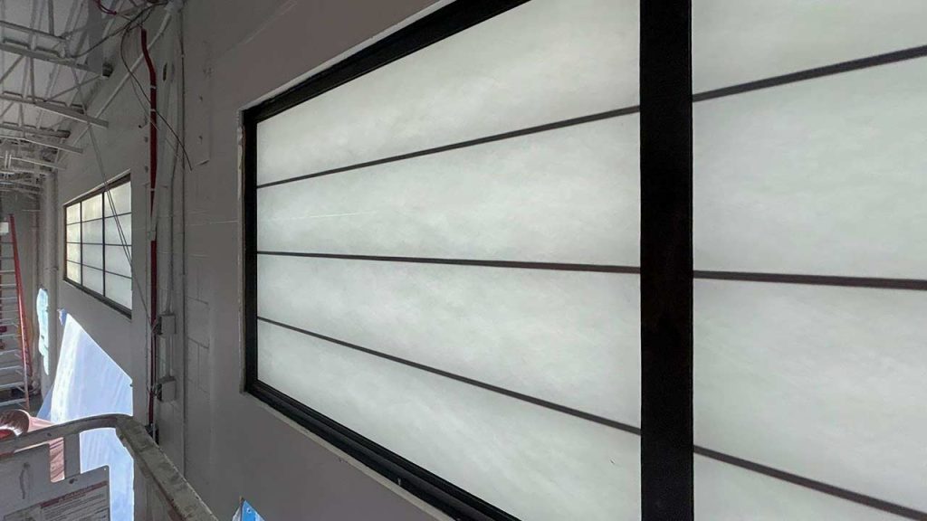 A white interior wall features two large rectangular wall panels with black frames and horizontal grilles. The scene is industrial with exposed wiring and piping.