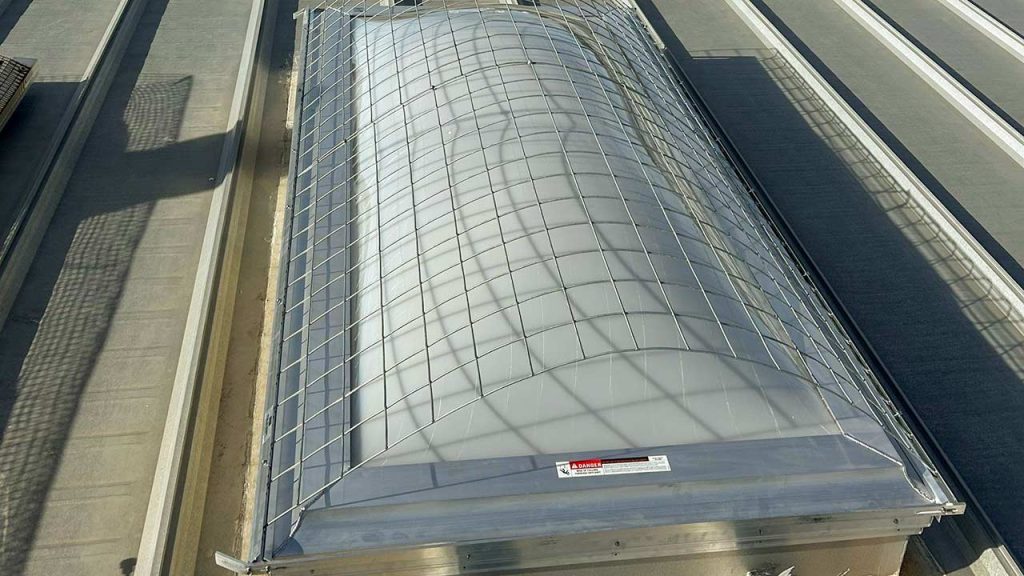 A large corrugated plastic skylight covered with a protective metal grid on an industrial rooftop. Sunlight casts grid shadows, creating a geometric pattern.