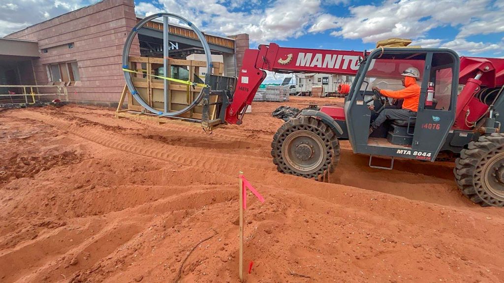 A red telescopic handler lifts a circular metal frame on a construction site. The ground is reddish-brown soil, with pink flags marking areas. The sky is partly cloudy.