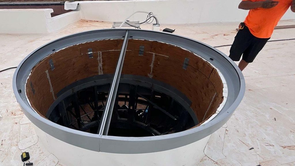 Rooftop installation of a large, circular open shaft with metal and wooden lining. A person in an orange shirt stands nearby, assessing the setup.