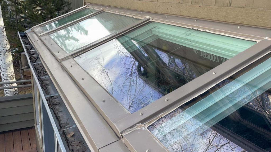 A close-up of a rooftop with glass skylight reflecting a clear sky and tree branches. The panels are framed by metal borders. The scene is calm and bright.