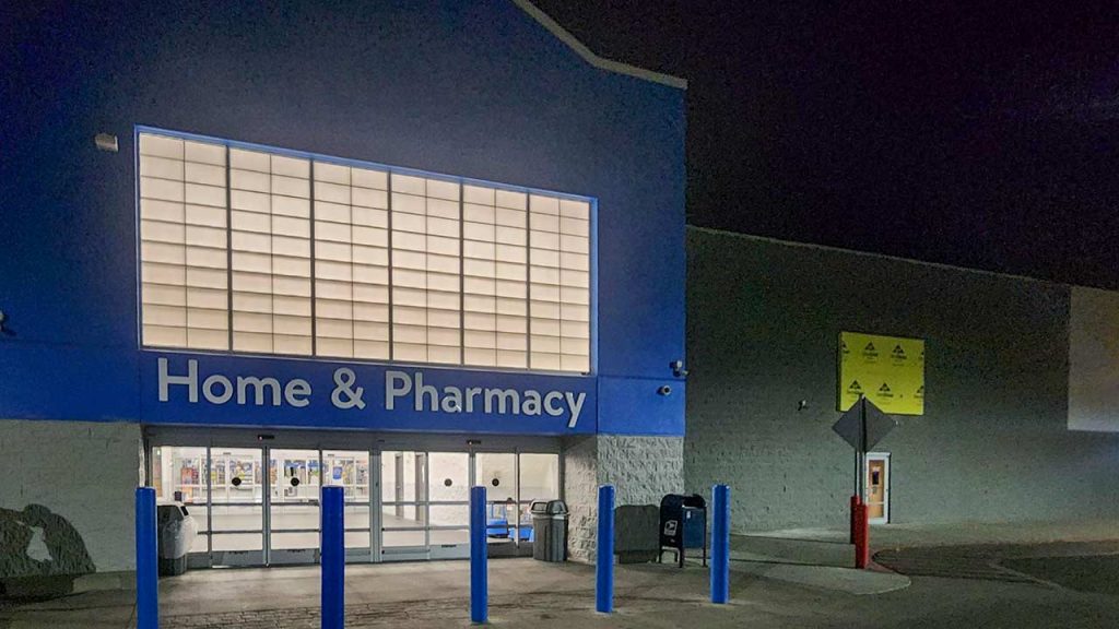 Night view of a closed retail store entrance labeled "Home & Pharmacy" with blue signage. The dimly lit scene shows tall blue posts and an empty parking lot.