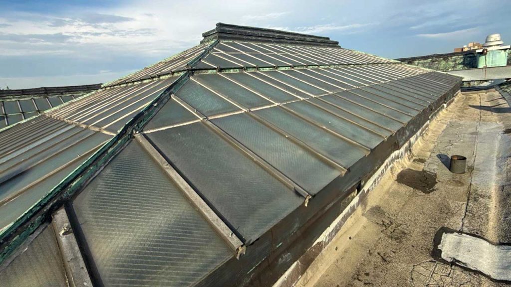 A historic glass and metal skylight on a rooftop under a blue sky, showing signs of aging with some rust, surrounded by a weathered surface.