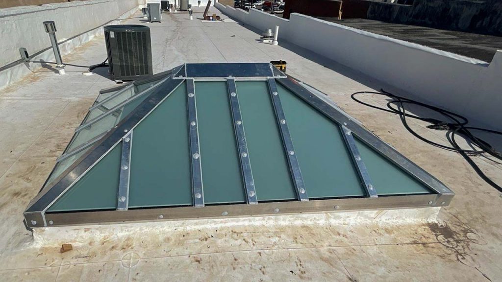 A glass pyramid-shaped skylight with metal framing sits on a flat rooftop, surrounded by HVAC units and tubing, under a clear blue sky.