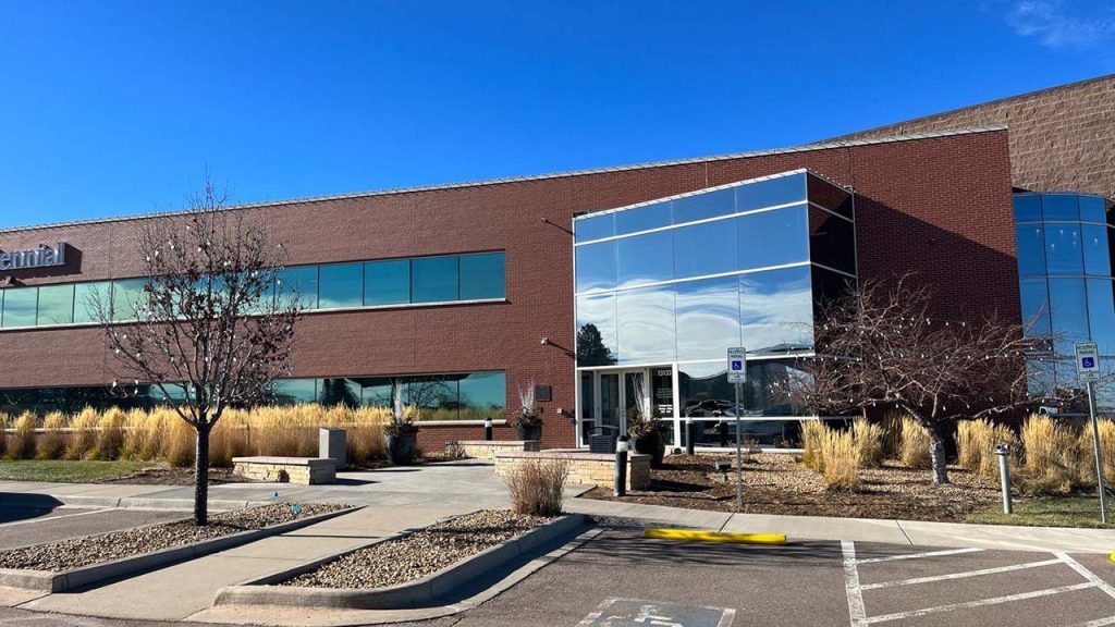 A modern brick office building with large reflective windows under a clear blue sky. Surrounding landscaping features dry grasses and leafless trees.