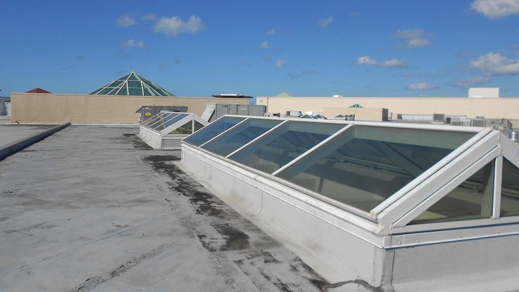 A flat rooftop under a clear sky with several large, ridge skylights. In the background, a green glass pyramid skylight is visible.