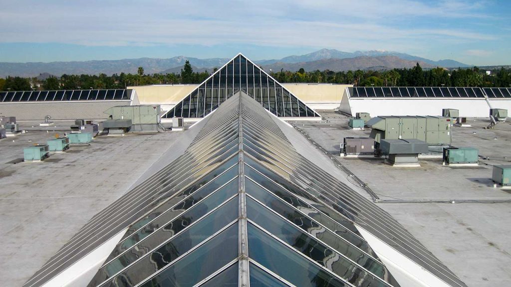 Rooftop with large glass skylights forming a triangular pattern, surrounded by air conditioning units. In the background, distant mountains and blue sky create a serene atmosphere.