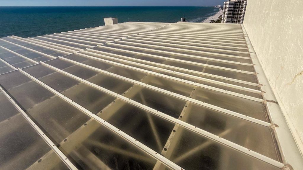 Rooftop with clear panels overlooking a vibrant blue ocean and a distant horizin. The scene conveys an airy, open feeling with a coastal vibe.