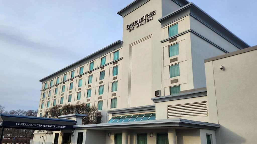 Exterior view of a DoubleTree by Hilton hotel, featuring a multi-story beige building with a single slope skylight and teal windows. Signs indicate a conference center entry.